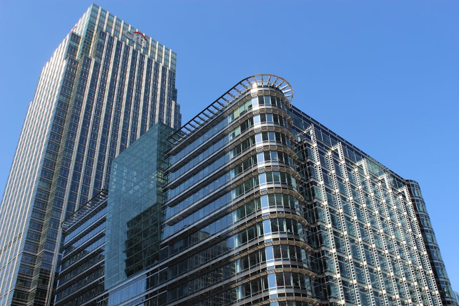 A modern multi-story office building with a curved corner, constructed primarily of glass panels framed by metal, reflects the surrounding cityscape on its sleek surface. The building's exterior featu